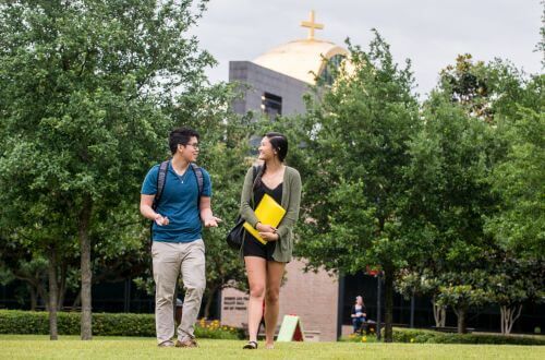 Two undergraduate students walking on campus at the University of St. Thomas in Houston, Texas with the Catholic Chapel of St. Basil visible in the background